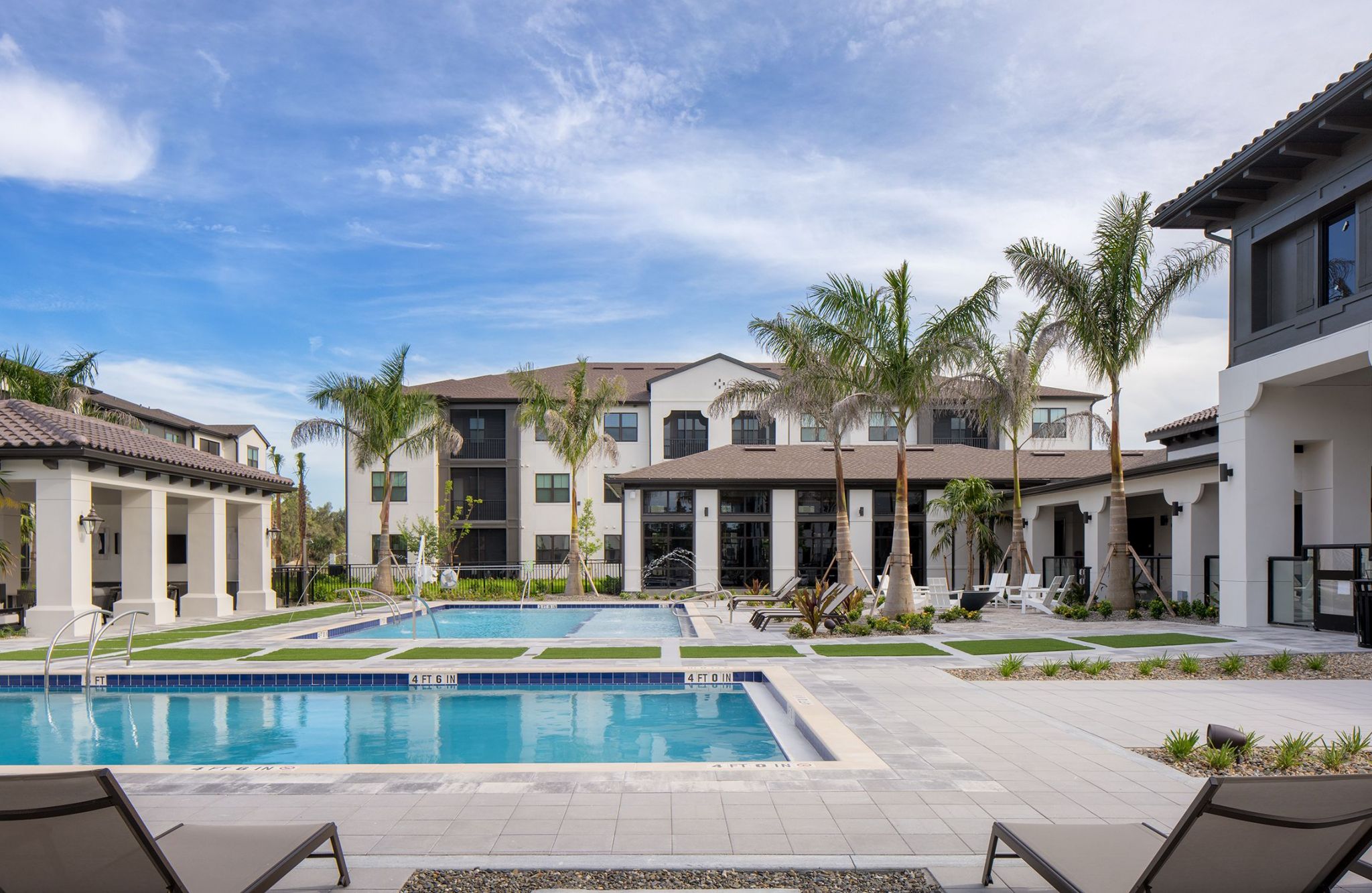 Modern apartment complex with a swimming pool, lounge chairs, and palm trees under a blue sky.