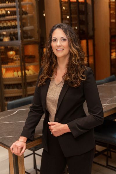 A woman with long curly hair in a black suit stands by a table in a room with wine racks.