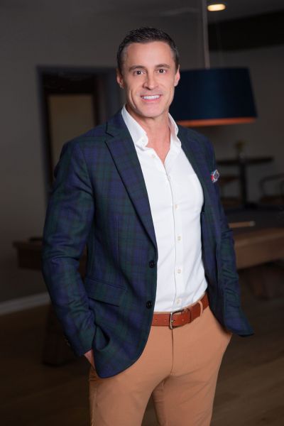 Man smiling in a plaid blazer and beige pants, standing indoors with his hands in his pockets.