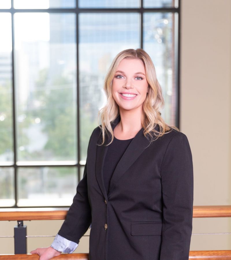 Person in a black suit standing by a railing, smiling, with large windows and a cityscape in the background.