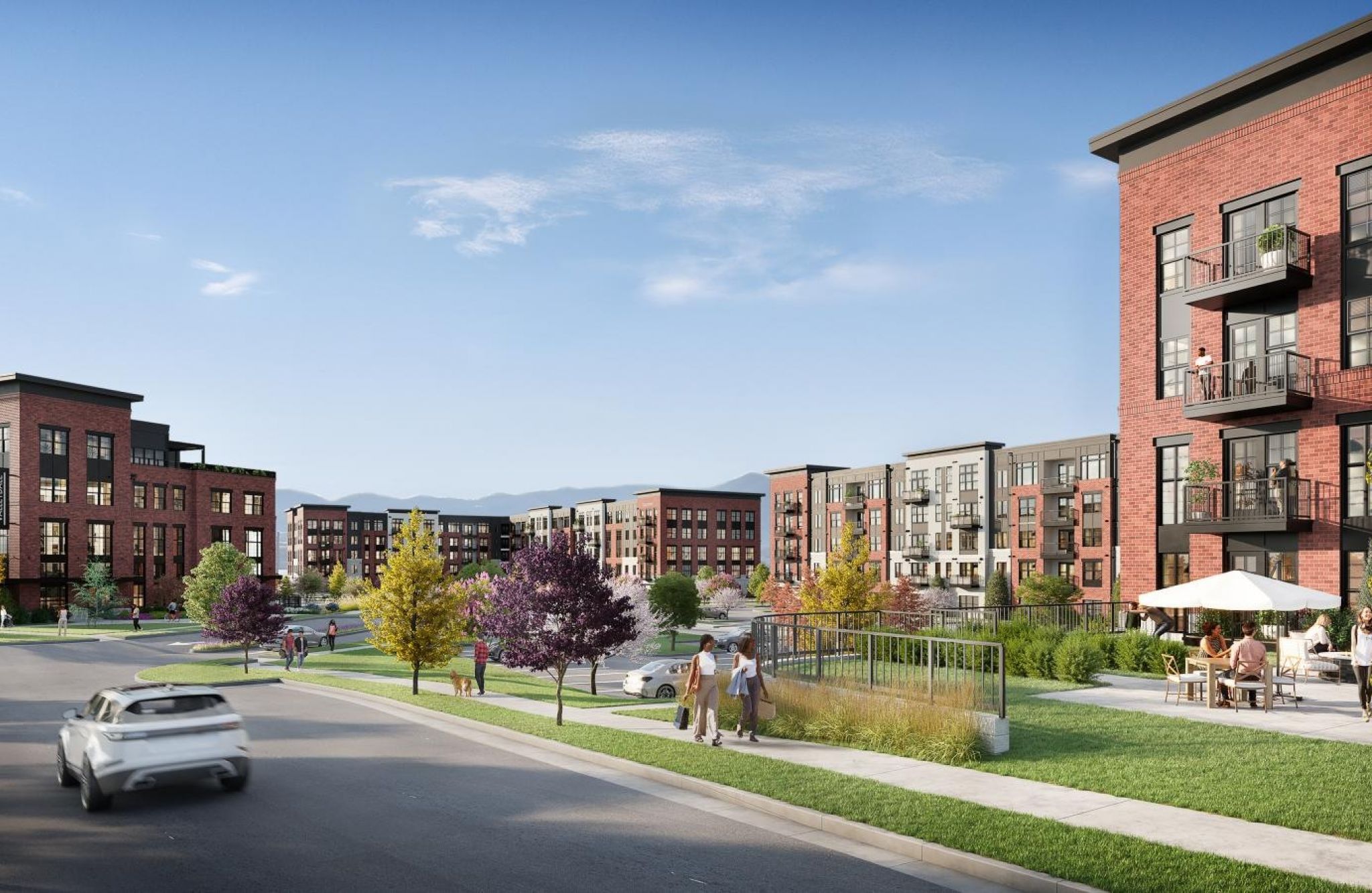 Modern apartment complex with red brick buildings, landscaped lawns, people walking, and a car on the road.