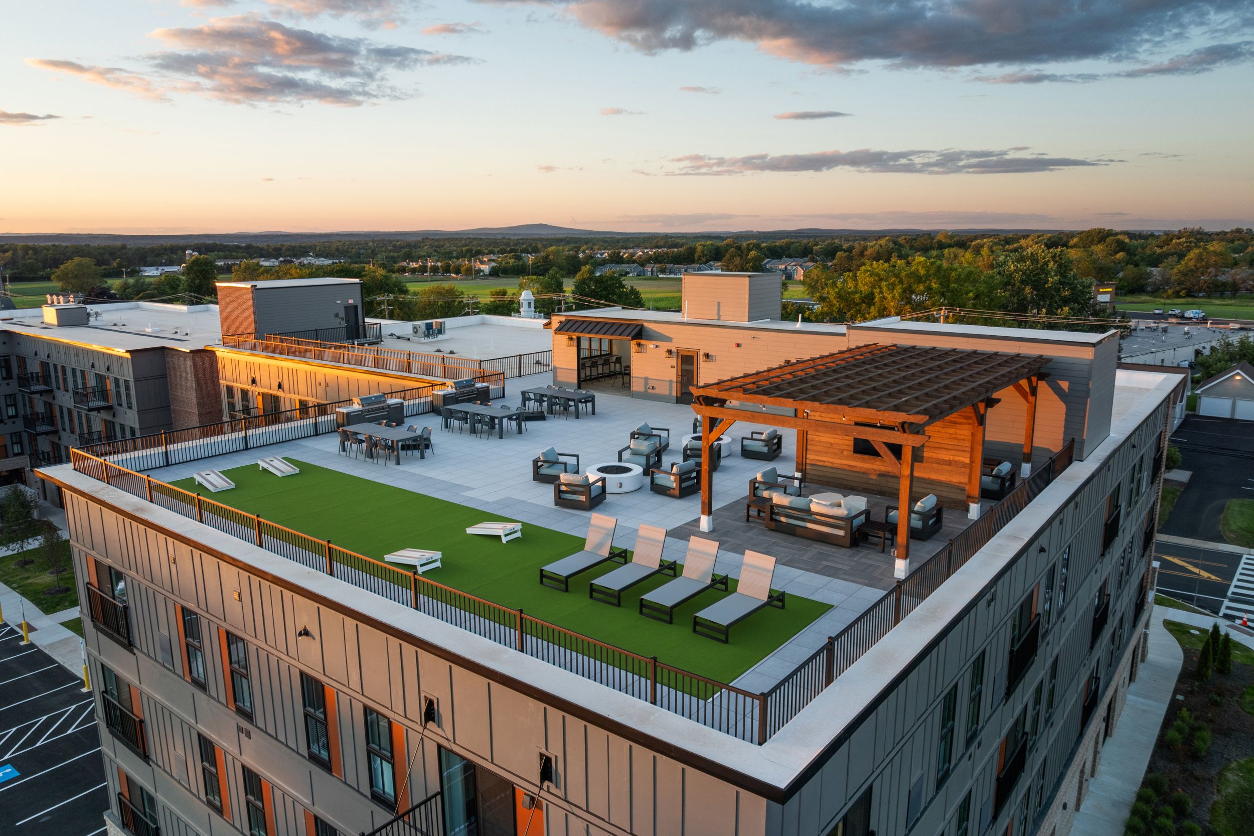 A modern apartment rooftop with lounge chairs, pergola, seating areas, and green turf, overlooking a suburban landscape at sunset.