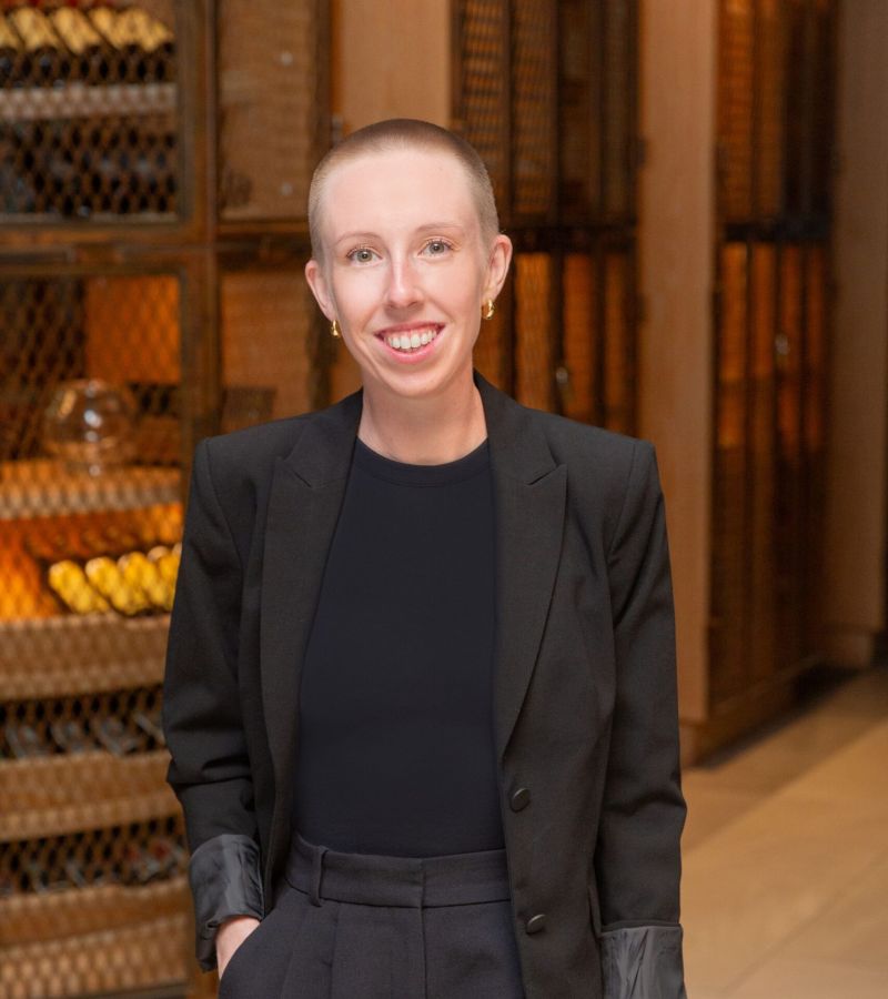 Person in a black suit smiling, standing indoors with hands in pockets, with wooden wine racks in the background.