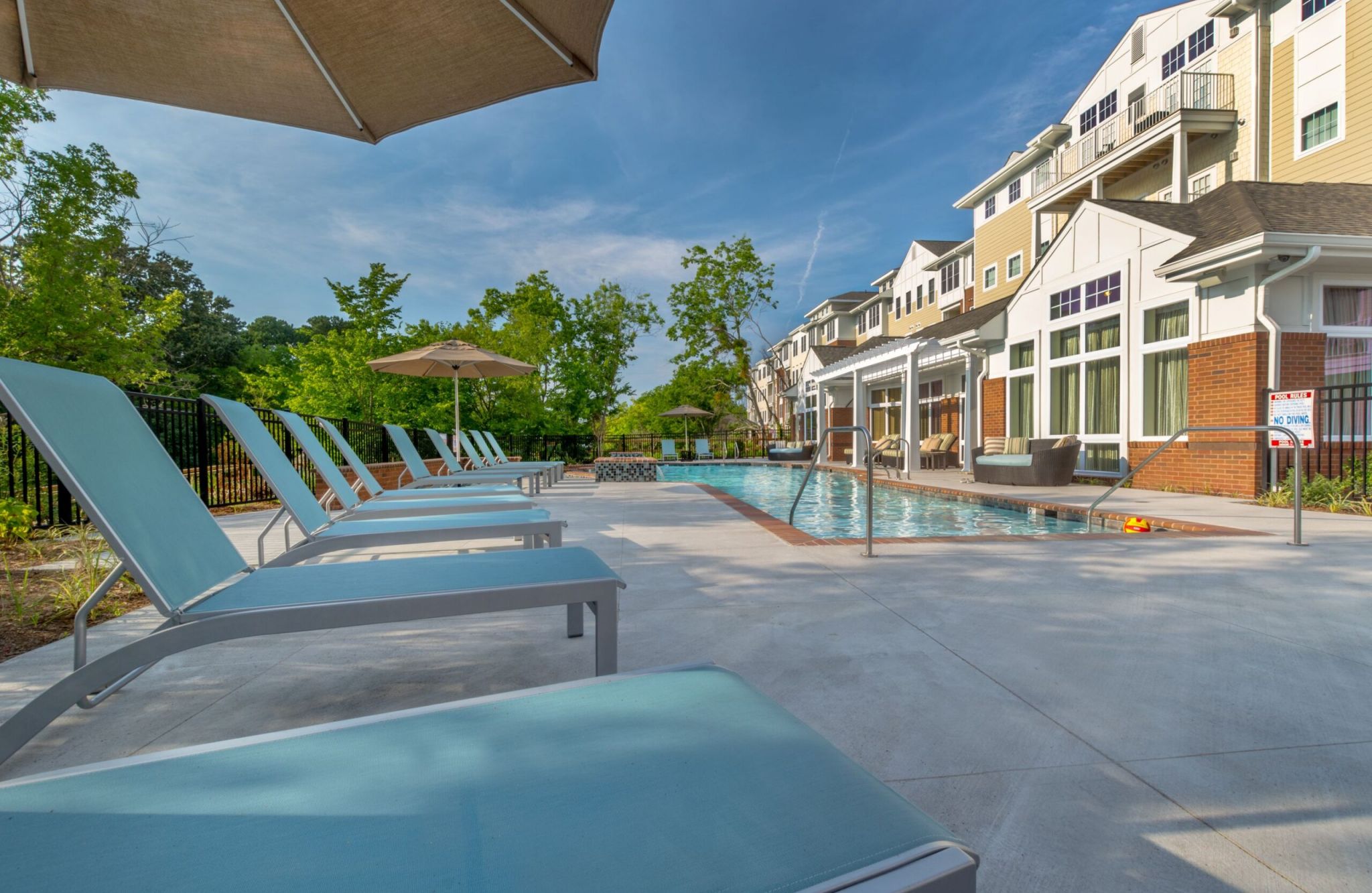 Poolside view with lounge chairs, umbrellas by Promenade Pointe, and a modern building in the background, surrounded by trees.