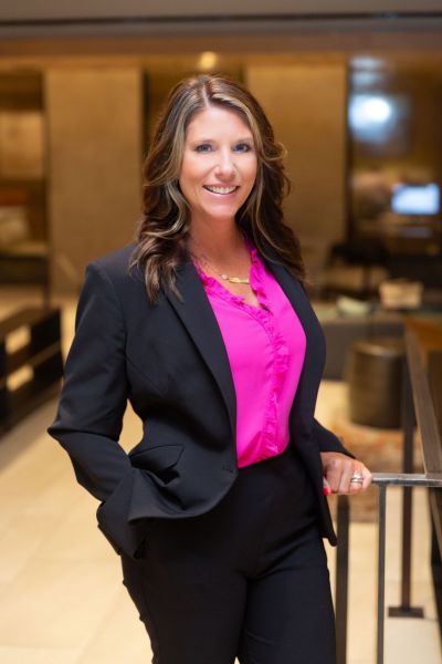 A woman in a black suit and pink blouse stands smiling in a modern office setting.