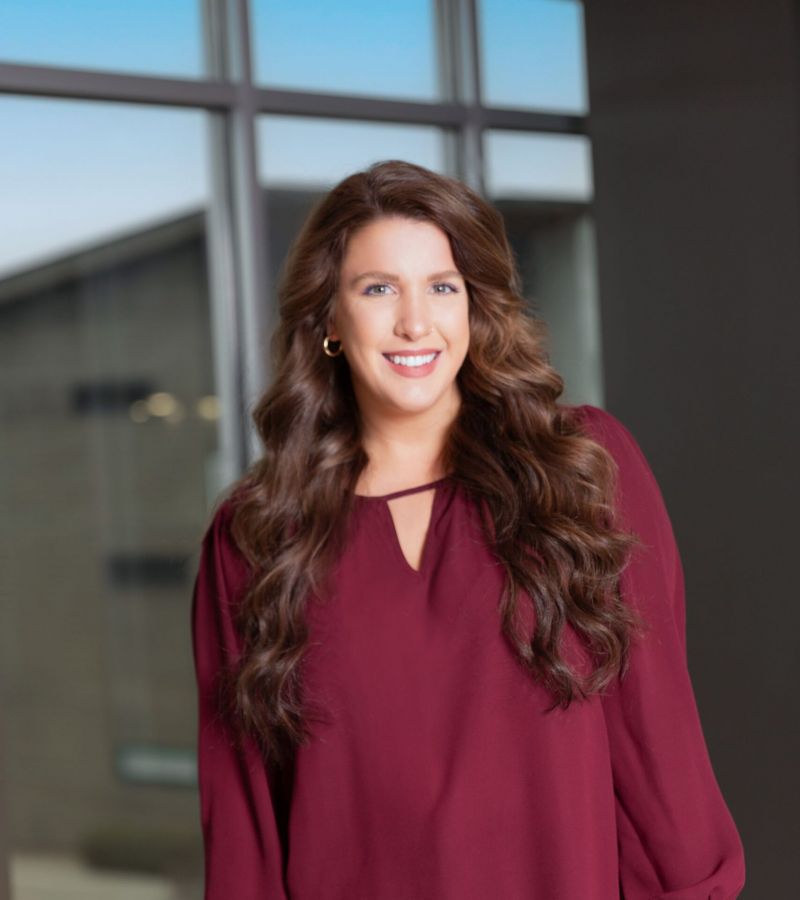 Smiling woman in a maroon blouse stands by a table in a modern office with large windows.