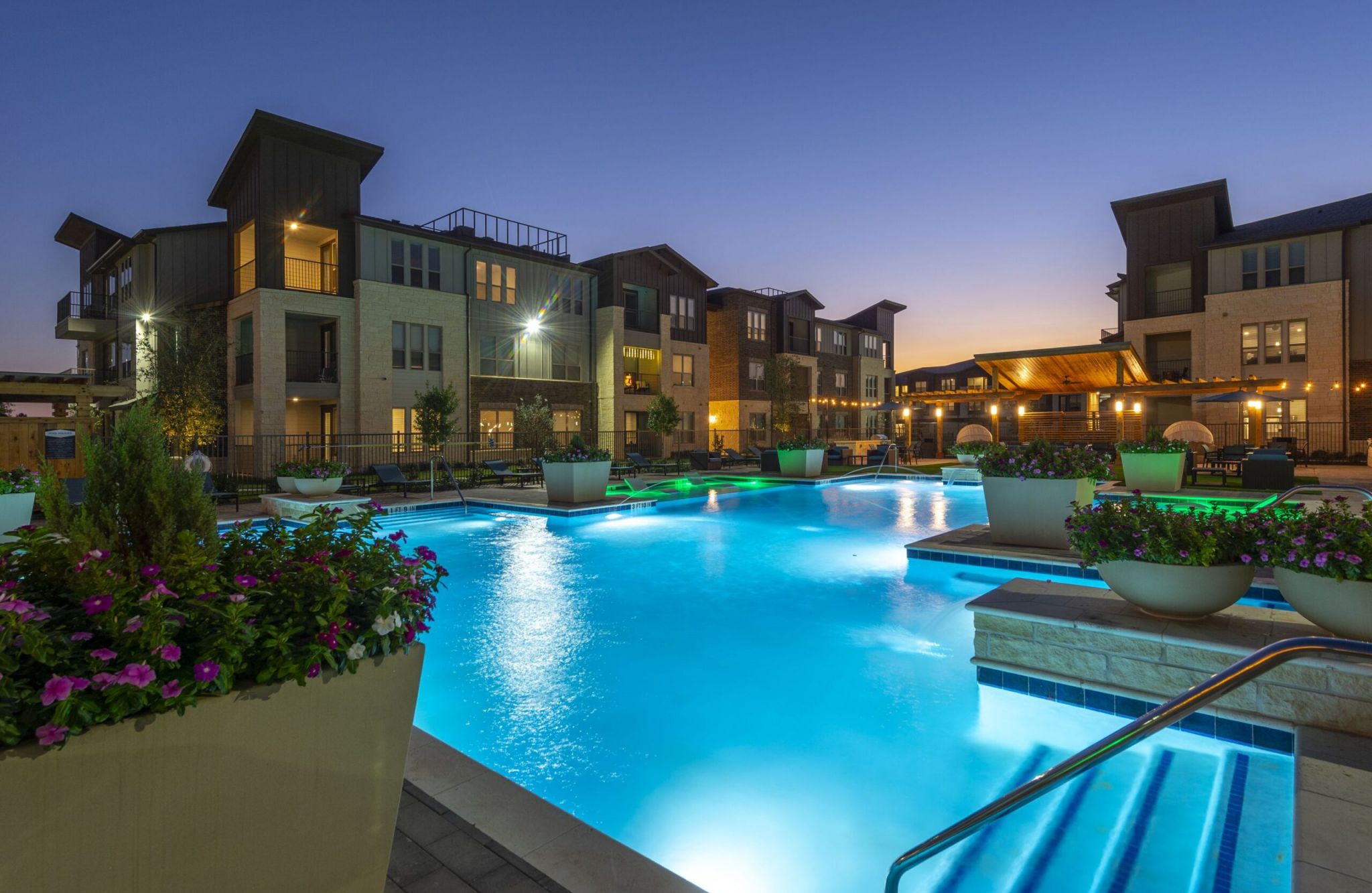 Illuminated pool with surrounding potted plants, nestled between Braxton Tammaron's modern apartment buildings at twilight.