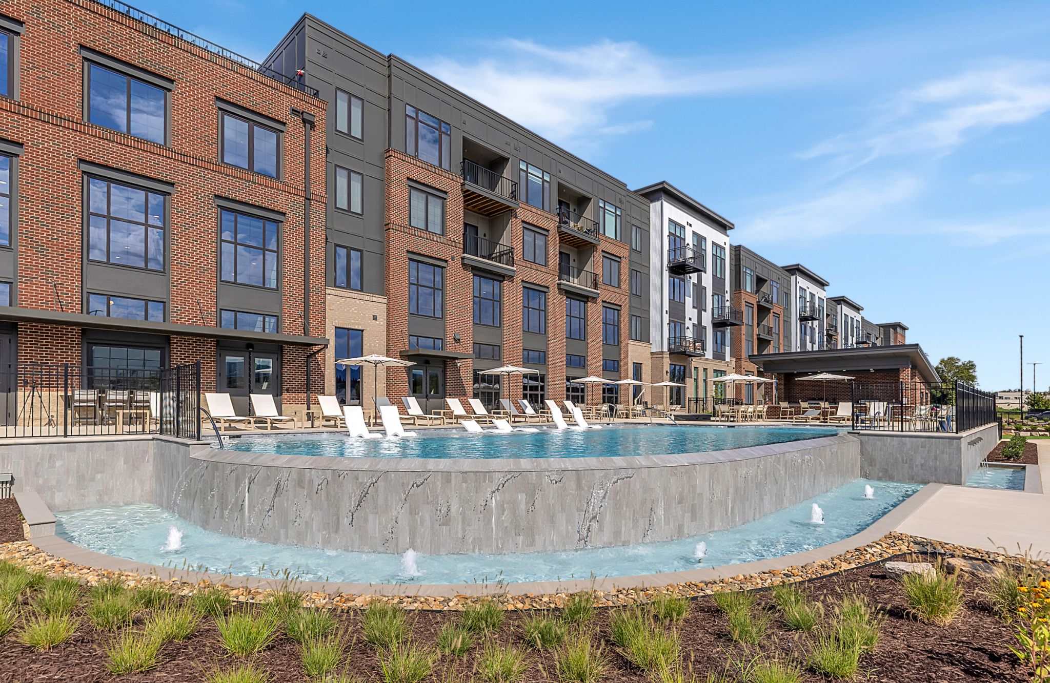 Modern apartment building with large windows, balconies, and an outdoor pool with lounge chairs and small fountains in front.