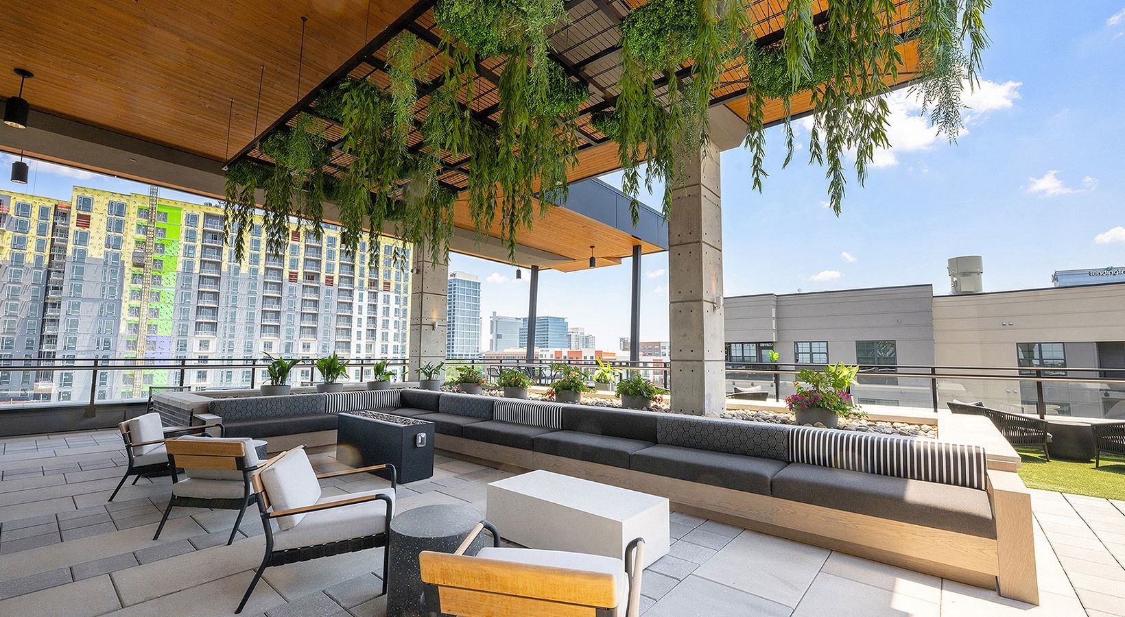 Modern rooftop patio with seating, hanging plants, and city views in the background under a clear blue sky.