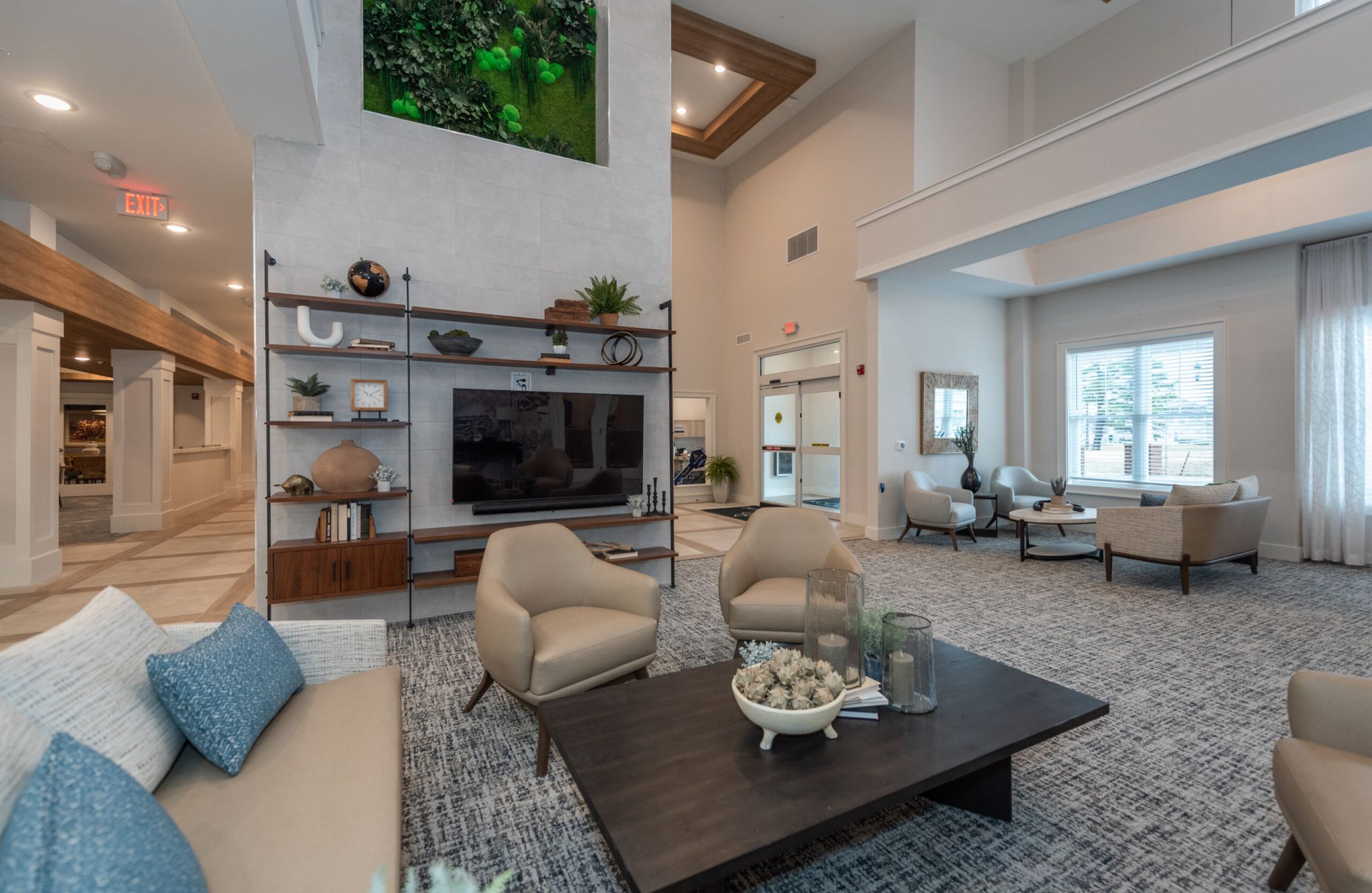Modern lobby at Acclaim at Greenbrier features beige chairs, coffee table, TV, large windows, shelves, and a lush green plant wall.