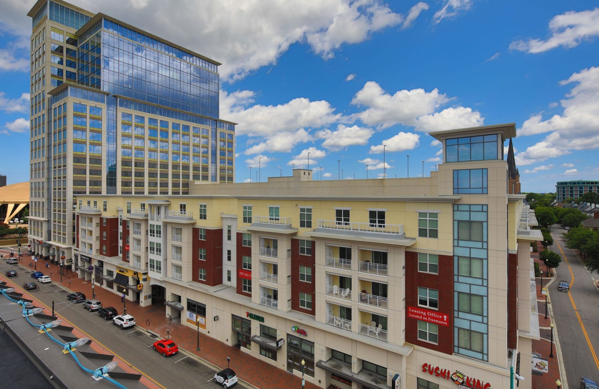 A street view of a modern downtown building with shops and apartments, under an attainable partly cloudy sky.