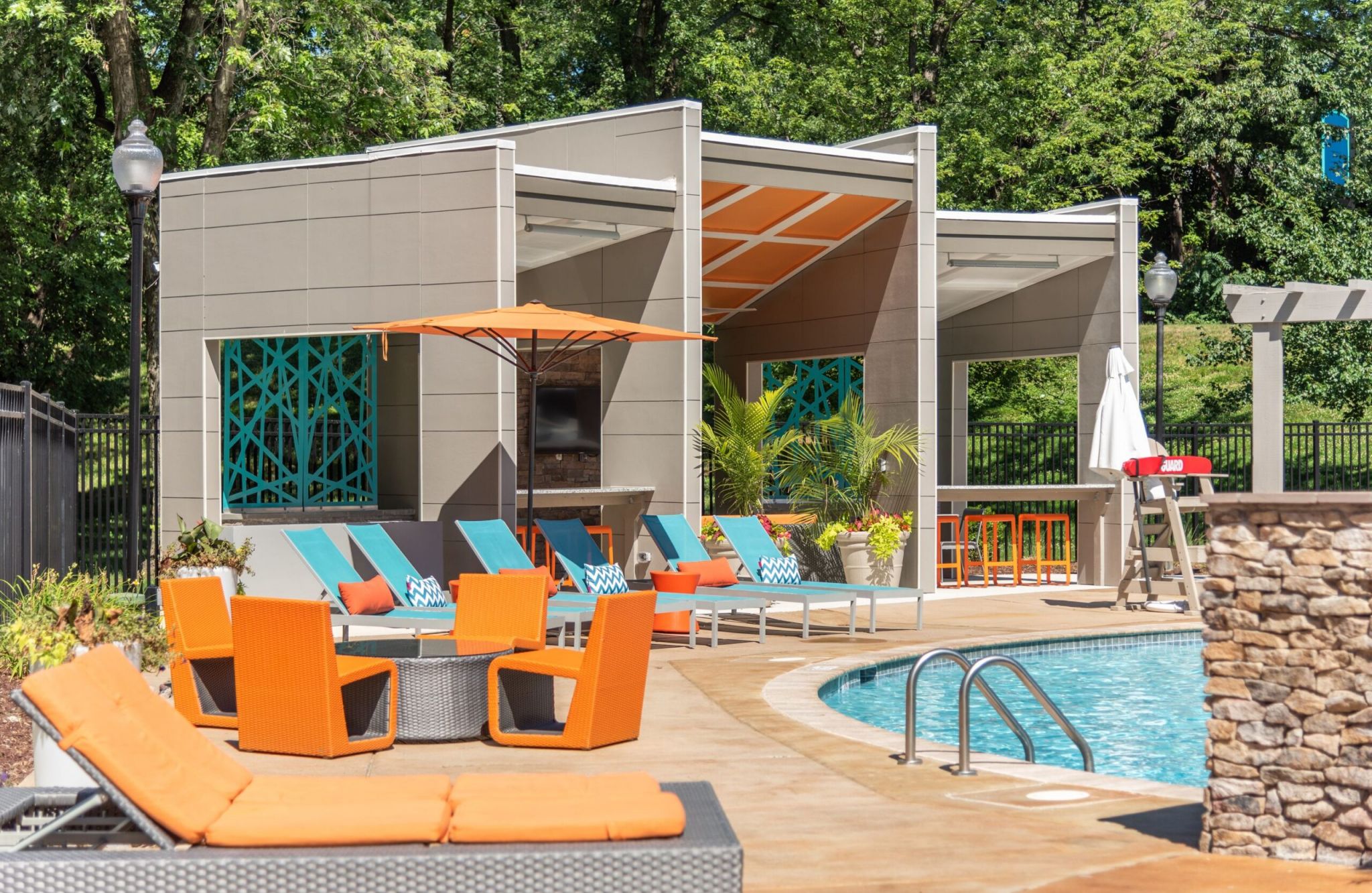 Outdoor pool area with orange lounge chairs, cabanas, and a modern building backdrop surrounded by trees.
