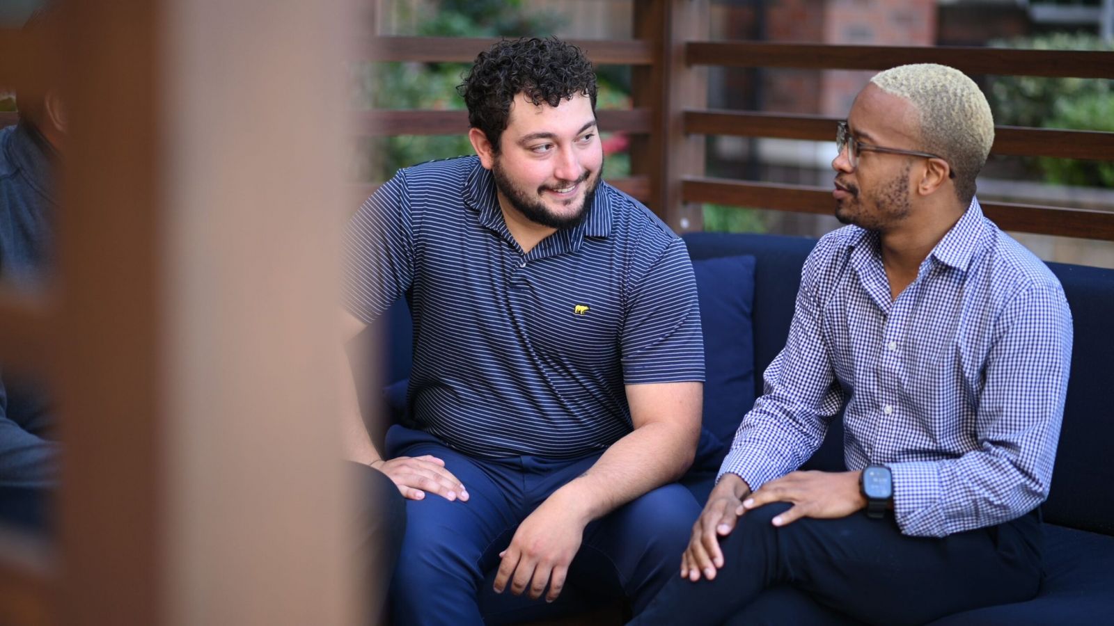 Two men seated on a blue couch outdoors, engaged in conversation, surrounded by wooden railings.
