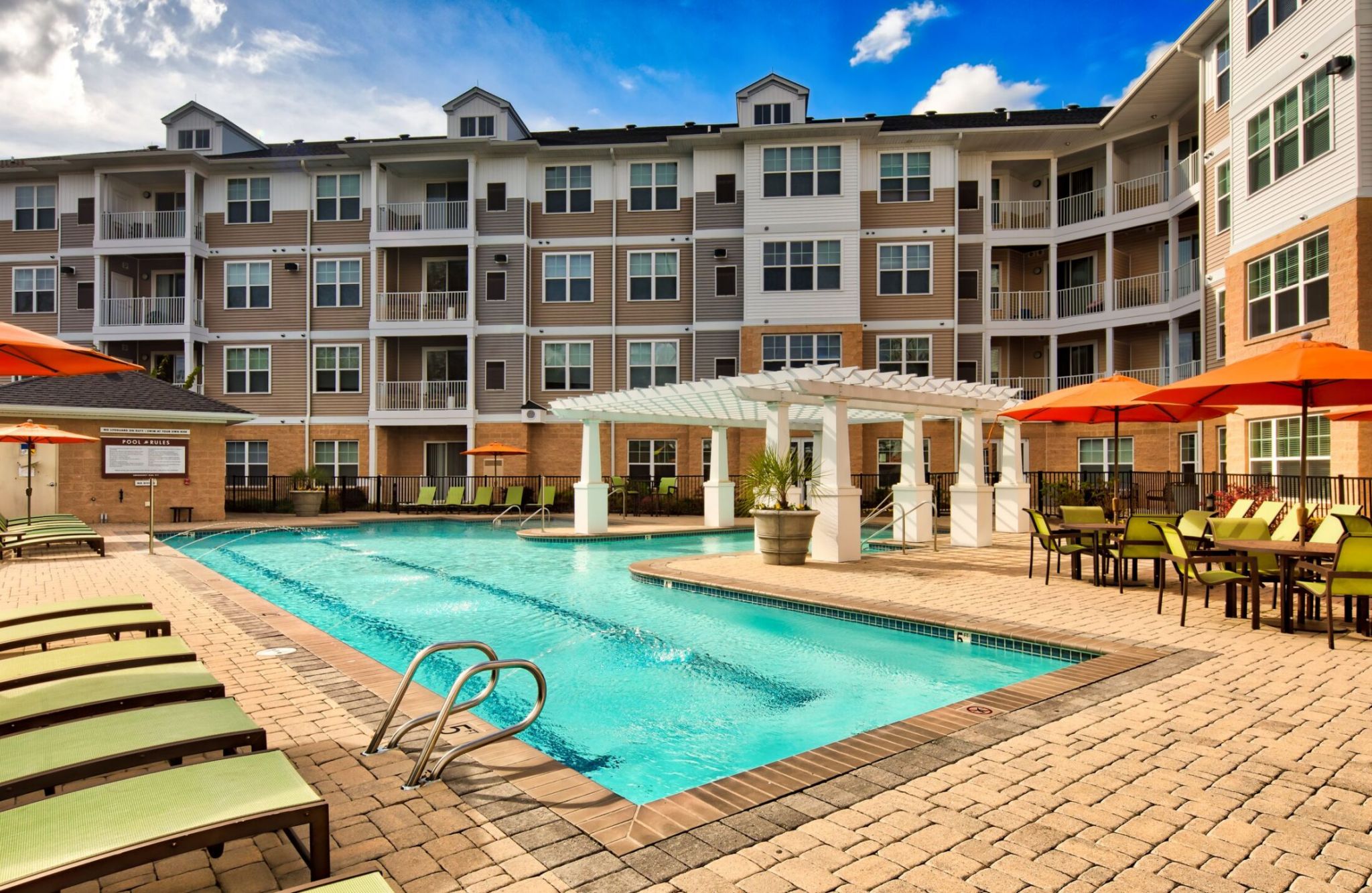 Apartment complex with pool, surrounded by lounge chairs and tables with orange umbrellas under a blue sky.