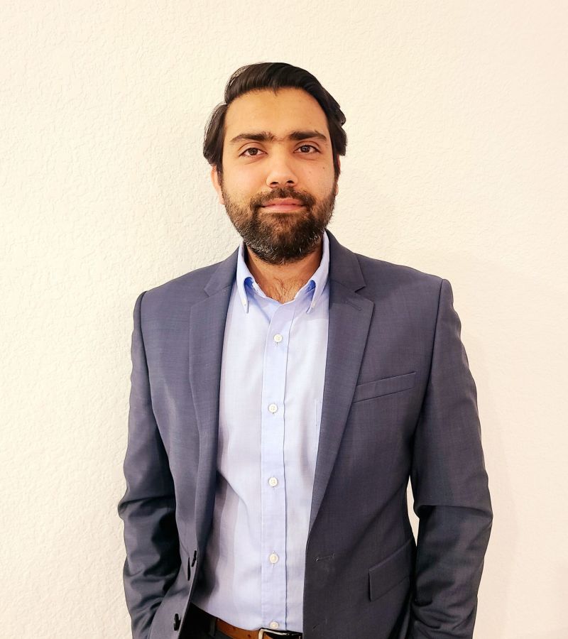 A man in a blue suit and light blue shirt stands in front of a white wall, looking at the camera.