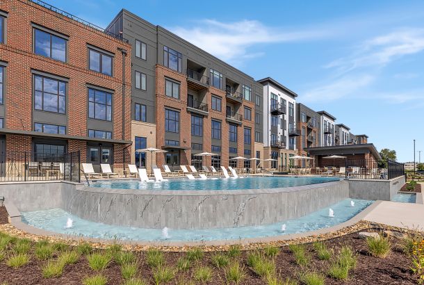 Modern apartment building with large windows, balconies, and an outdoor pool with lounge chairs and small fountains in front.