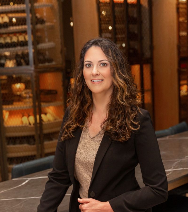 A woman with long curly hair in a black suit stands by a table in a room with wine racks.