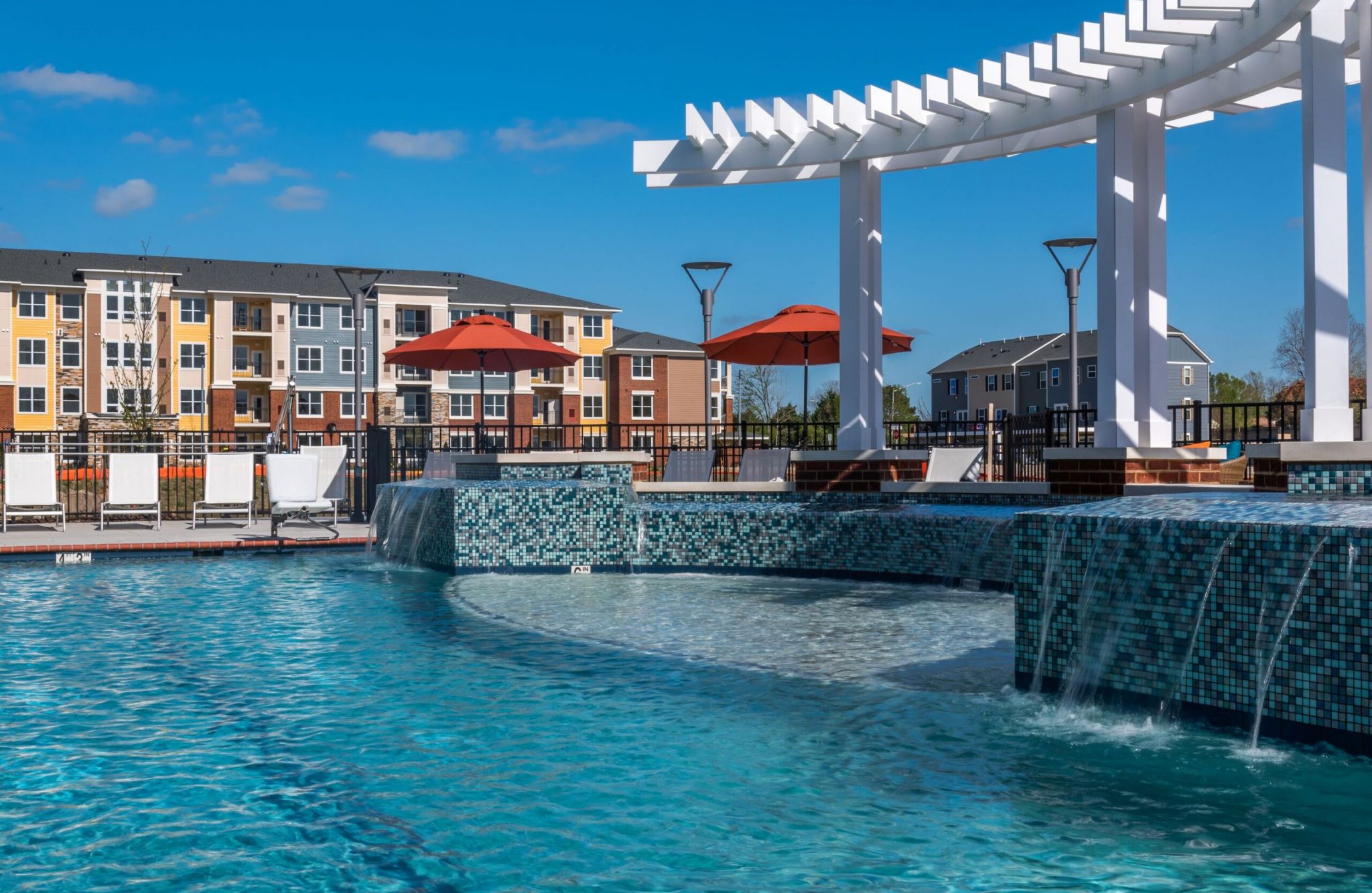 Infinity pool with a white pergola, lounge chairs, and Centerville Crossing's brick apartments in the background on a sunny day.