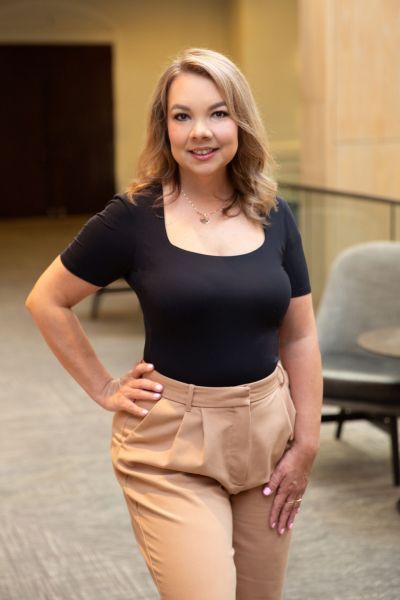 Smiling woman in a black top and beige pants stands in a modern indoor setting with a hand on her hip.