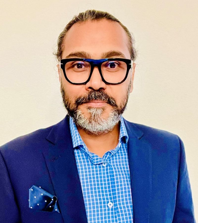 A man with glasses and a beard, Sharouq Shahzad, wears a blue blazer and checked shirt against a light background.