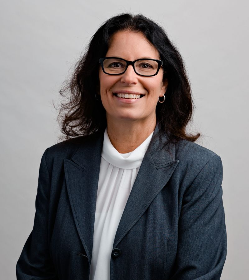 Woman with long dark hair, wearing glasses and a gray suit jacket, smiling in front of a plain background.