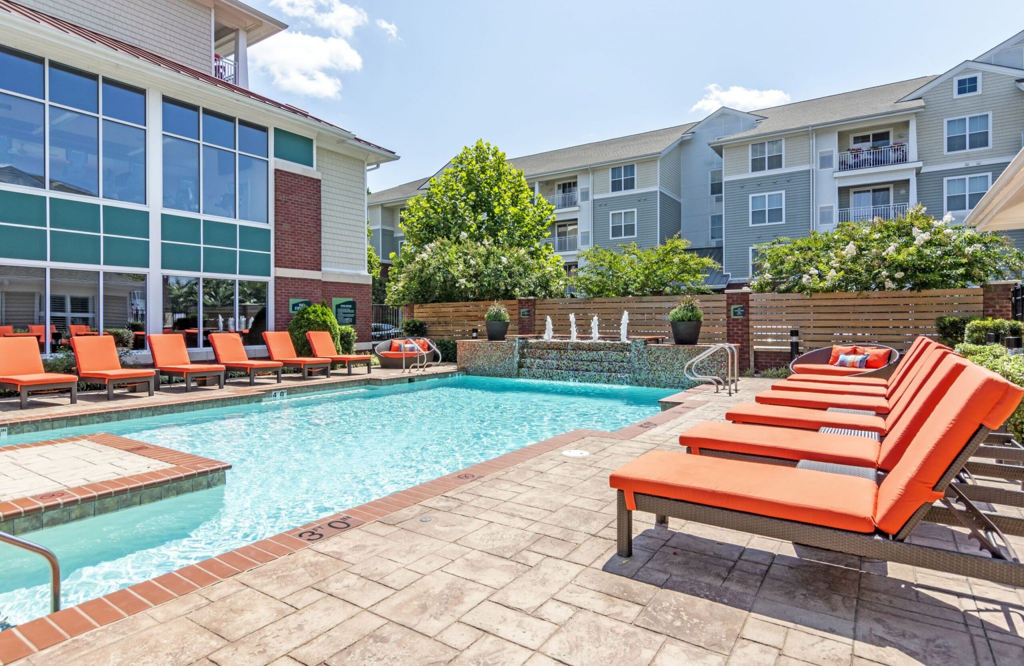 Outdoor pool area at Carlton, featuring red lounge chairs, a small fountain, and modern apartment buildings in the background.