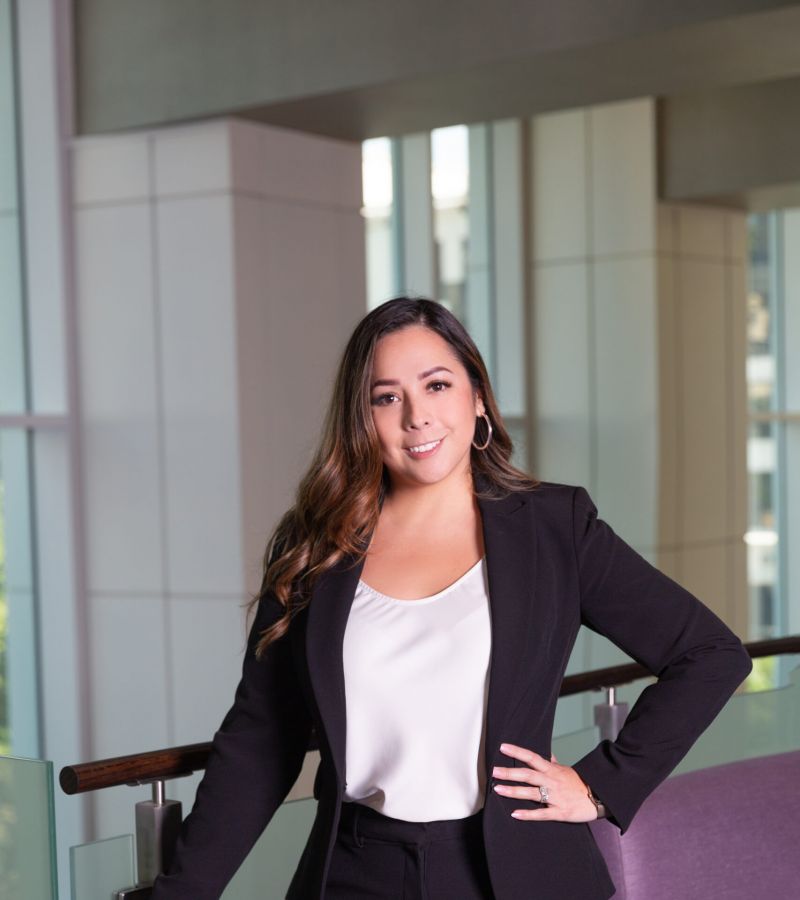 Woman in a black suit and white blouse stands confidently by a railing in a modern office building.