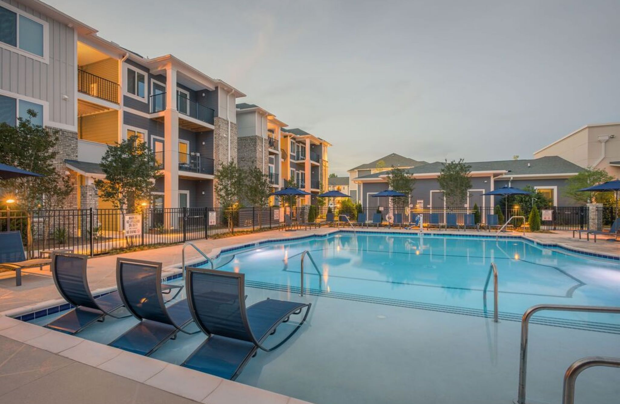 Rivergate apartment complex boasts a tranquil pool and lounge chairs, nestled among modern buildings at dusk—a true haven.