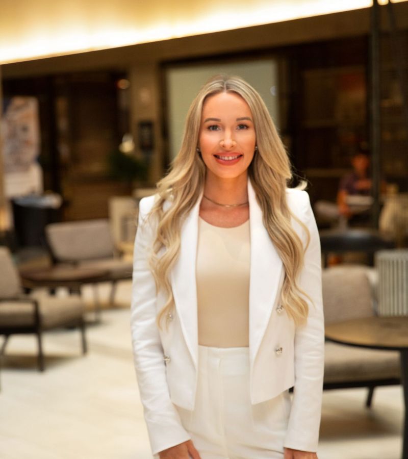 A woman in a white suit stands in a modern, well-lit indoor setting with tables and chairs in the background.