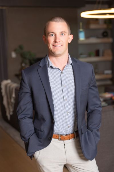 A man in a suit stands in a modern room with hands in pockets, smiling at the camera.