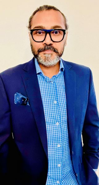 A man with glasses and a beard, Sharouq Shahzad, wears a blue blazer and checked shirt against a light background.
