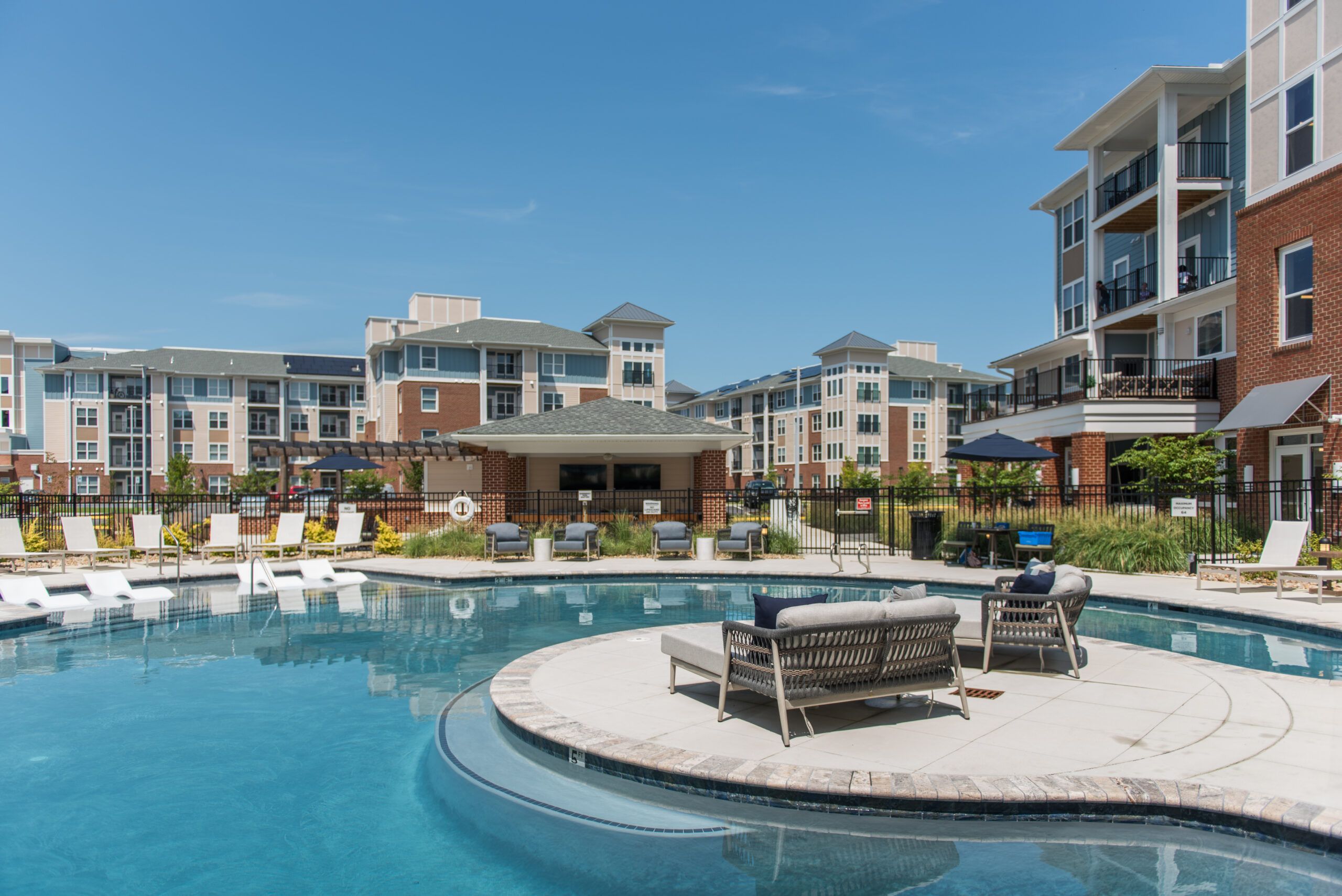 Modern apartment complex with a large outdoor pool, lounge chairs, and patio seating under a clear blue sky.