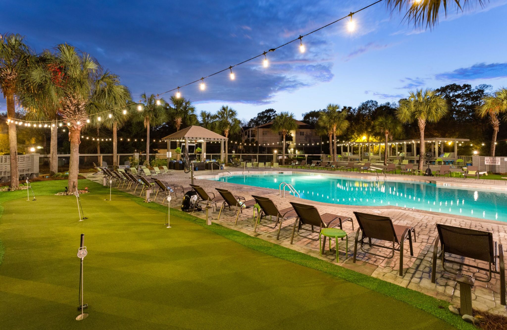 Riverland Woods' outdoor pool area at dusk, with string lights, lounge chairs, and palm trees, features a mini-golf area in the foreground.