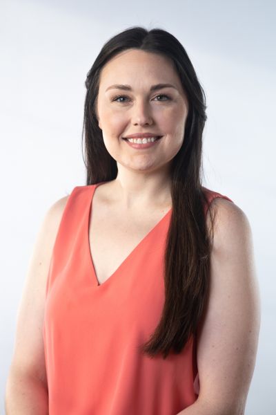 A woman with long brown hair smiles while wearing a sleeveless coral top against a light background.