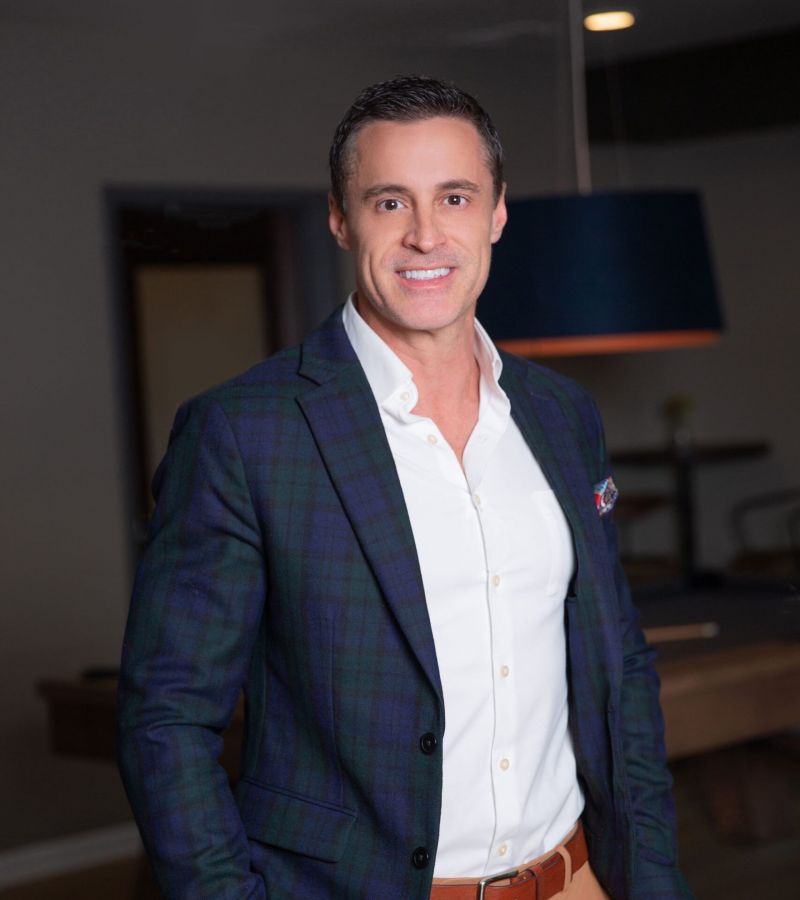 Man smiling in a plaid blazer and beige pants, standing indoors with his hands in his pockets.