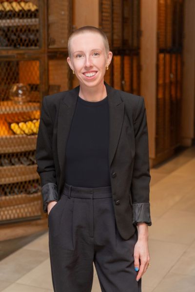 Person in a black suit smiling, standing indoors with hands in pockets, with wooden wine racks in the background.
