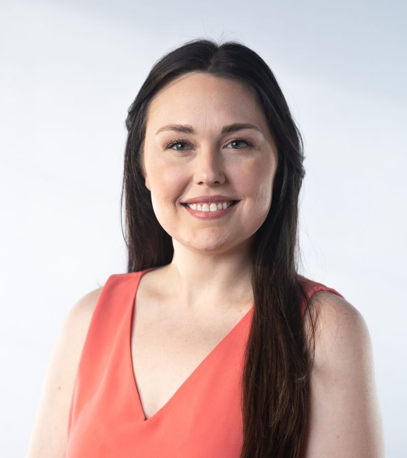 A woman with long brown hair smiles while wearing a sleeveless coral top against a light background.