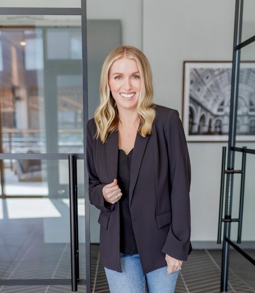 Woman with blonde hair in a black blazer and jeans smiles while standing in a modern office space at Summer Waters.
