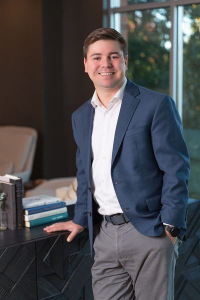 Person in a blue blazer smiles while leaning on a table with books in a well-lit room.