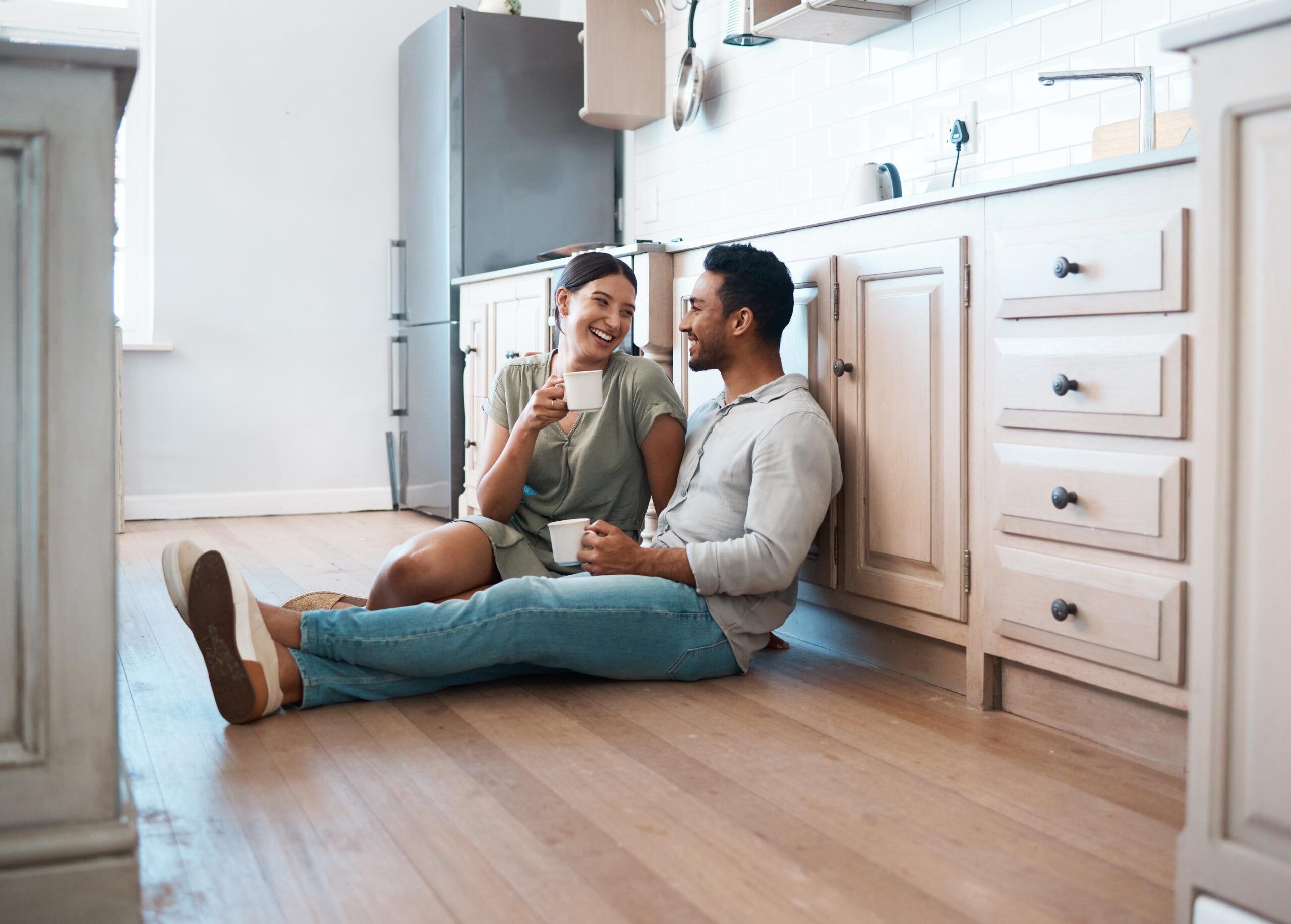 A couple sits on a kitchen floor, smiling and holding mugs, as natural light streams into their Vest Residential home.