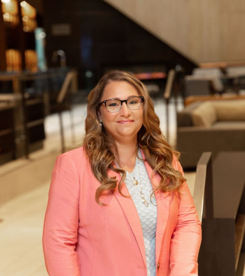 Person in glasses wearing a pink blazer stands in a modern lobby.
