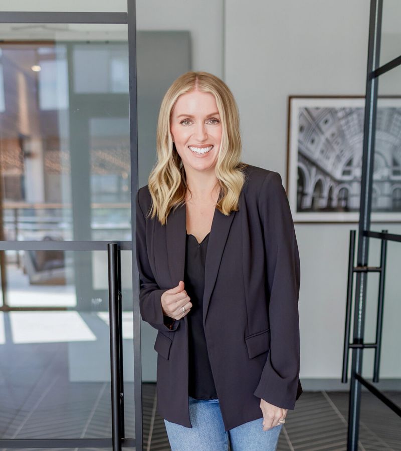 Woman with blonde hair in a black blazer and jeans smiles while standing in a modern office space at Summer Waters.