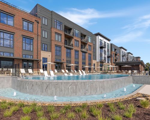 Modern apartment building with large windows, balconies, and an outdoor pool with lounge chairs and small fountains in front.