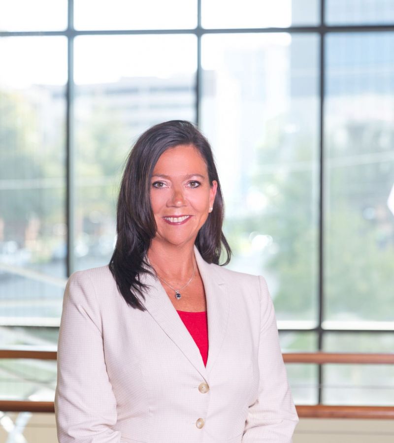 Woman in a cream suit standing indoors with large windows and trees in the background.
