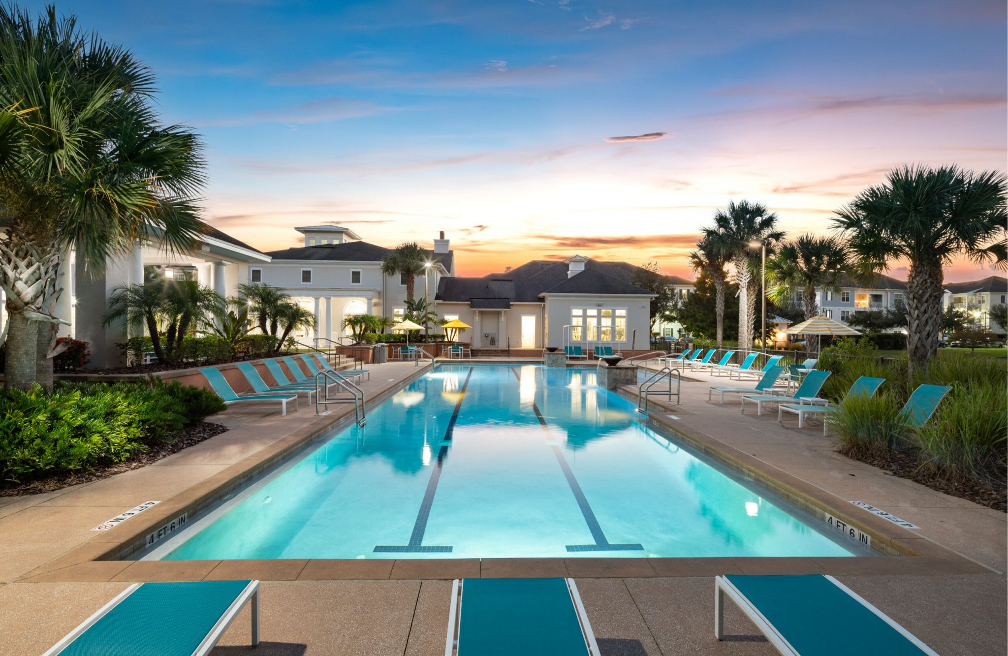 Rectangular outdoor pool at Evander Square with turquoise lounge chairs, palm trees, and buildings in the background at sunset.
