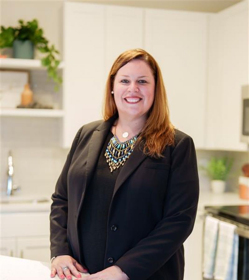 Smiling woman in a black suit, Ashley Dugger, stands in a modern kitchen with white cabinets and plants.