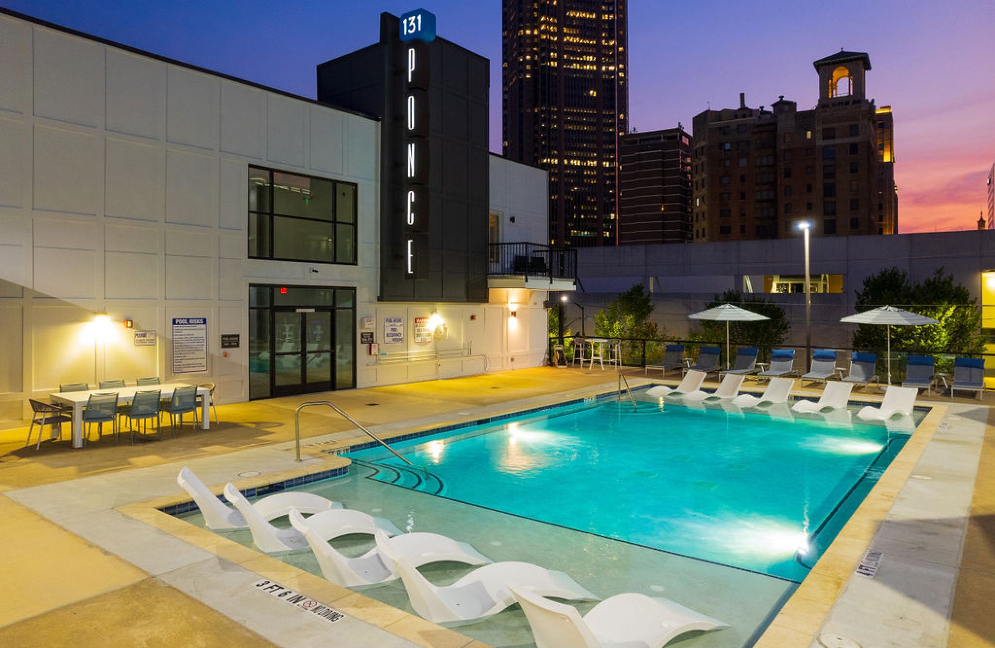 Rooftop pool with lounge chairs and tables, surrounded by city buildings at sunset.