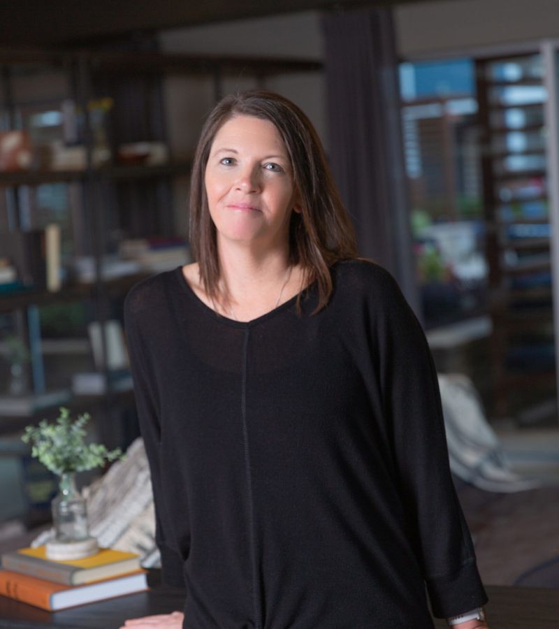 A woman in a black top and gray pants leans on a wooden table, with shelves and books in the background.