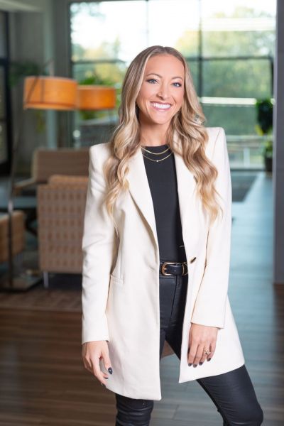 Smiling person in a white blazer and black outfit standing indoors with soft lighting and modern decor.