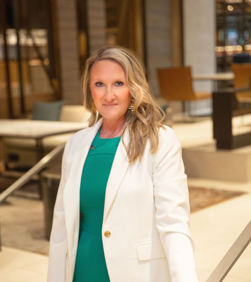 Woman in white blazer and green outfit standing indoors near stairs, looking at the camera.